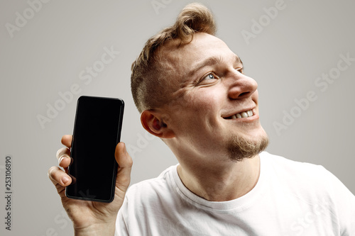Young handsome man showing smartphone screen over gray background with a surprise face. Human emotions, facial expression concept