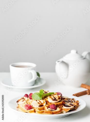Belgian curd waffles with raspberries, banana, chocolate syrup. Breakfast with tea on white background, side view, vertical
