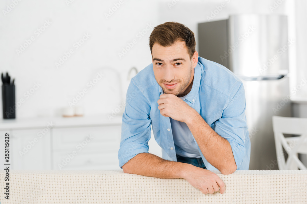 handsome smiling man propping chin with hand, leaning on sofa and looking at camera at home