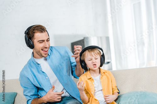 father with preschooler son in headphones listening music, gesturing with hands and playing imaginary guitars at home