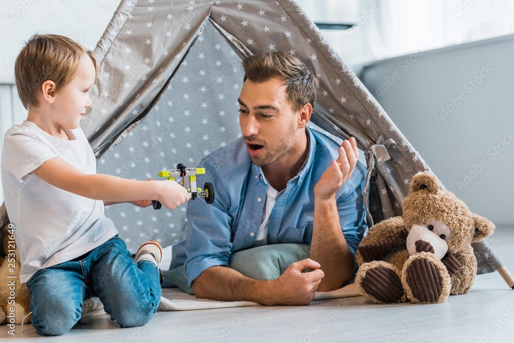 surprised father looking at preschooler son playing with toy car under wigwam at home