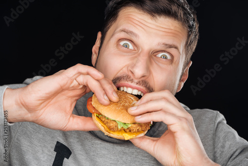 emotional man eating tasty chicken burger while looking at camera isolated on black