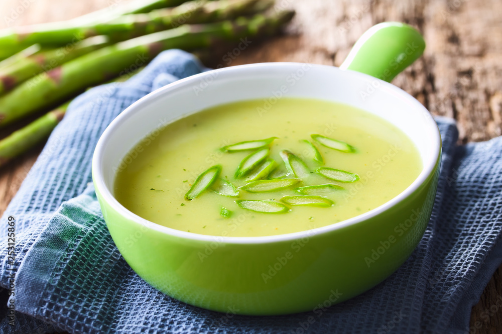 Cream of green asparagus soup in bowl, garnished with sliced asparagus on top, fresh asparagus in the back (Selective Focus, Focus in the middle of the asparagus slices on the soup) Cream of green asparagus soup in bowl, garnished with sliced asparagus on top, fresh asparagus in the back (Selective Focus, Focus in the middle of the asparagus slices on the soup)