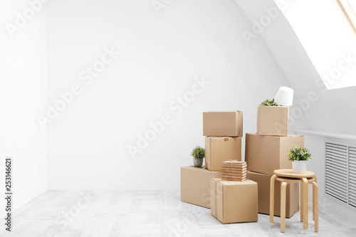 Moving to a new home. Belongings in cardboard boxes, books and green plants in pots stand on the gray floor against the background of a white wall. © spaskov