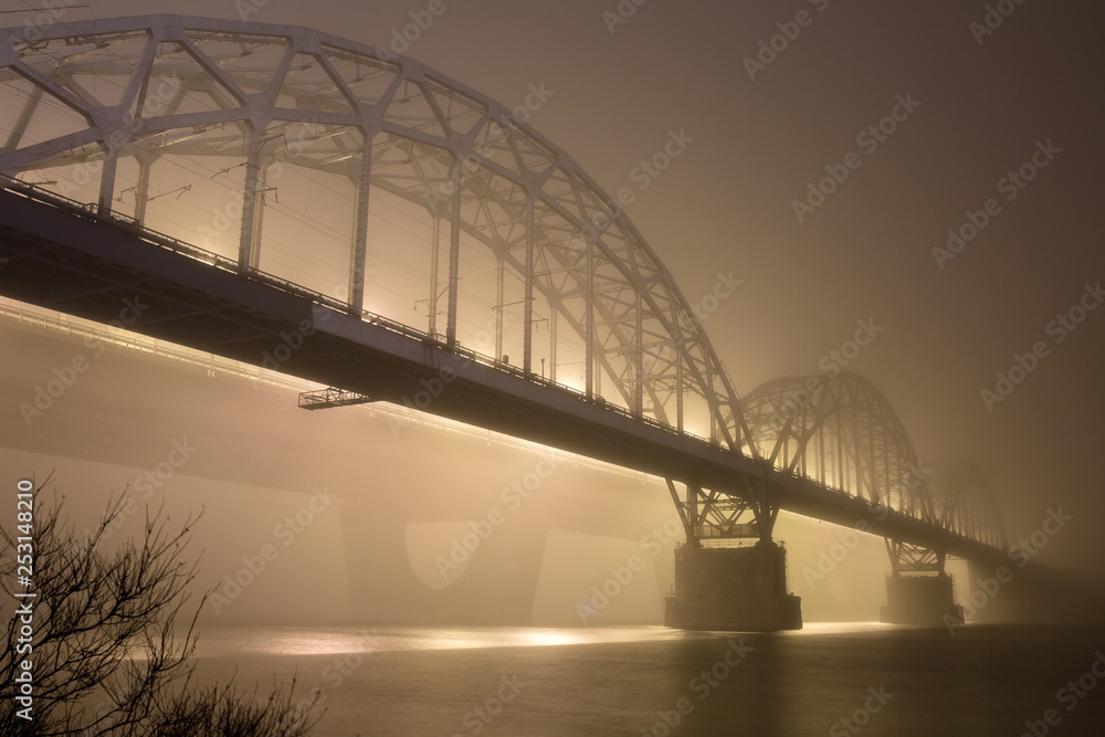 Obraz premium A mysterious evening fog above the river in big city. Bridge in the mist, cold weather scenery. Soft, blurry, misty look. Colorful, mystic industrial cityscape.