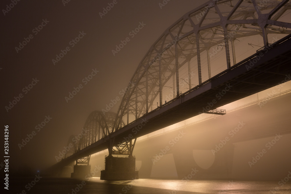 Obraz premium A mysterious evening fog above the river in big city. Bridge in the mist, cold weather scenery. Soft, blurry, misty look. Colorful, mystic industrial cityscape.