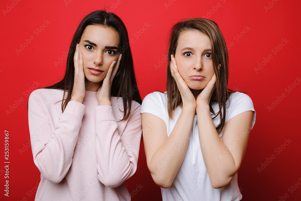 Two young pretty woman staring astonished into camera touching her cheeks with hands in front of a red background. Two young pretty woman staring astonished into camera touching her cheeks with hands in front of a red background.