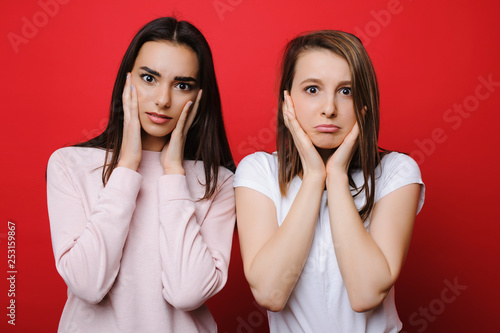 Two young pretty woman staring astonished into camera touching her cheeks with hands in front of a red background.