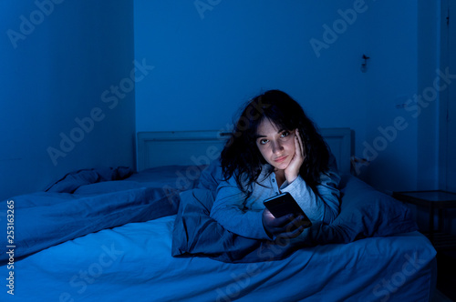 Attractive latin woman addicted to mobile phone and internet late at nigh in bed looking sleepless