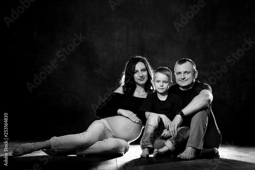 Happy family: pregnant mother with a tummy, father and son are sitting on a black background