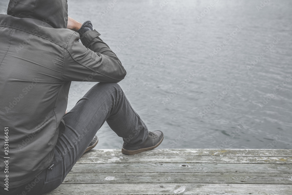 Young man sitting on edge of footbridge at lake. Suffering from life difficulties, broken heart or psychological problems. Dark, gloomy thoughts. Social isolation. Cool autumn time. Young man sitting on edge of footbridge at lake. Suffering from life difficulties, broken heart or psychological problems. Dark, gloomy thoughts. Social isolation. Cool autumn time.