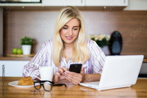 woman in pajamas drinking coffee and using smartphone and laptop in the kitchen