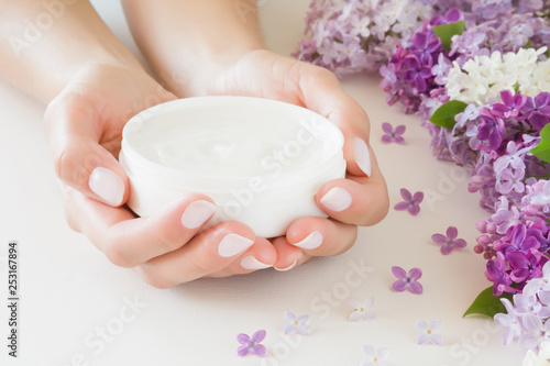 Young, perfect, groomed woman's hands holding moisturizing cream. Care about nails and clean, soft, smooth skin. Beautiful branches of fresh, colorful lilac flowers. Front view. Close up.