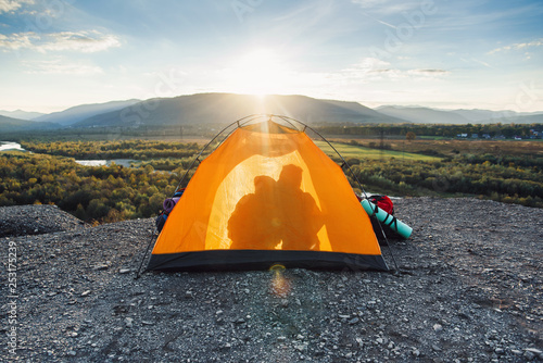 The concept of travel  tourism and relationships. Silhouette of a young man and his girlfriend who kisses each other in a orange tent on the top of the mountain at sunset.