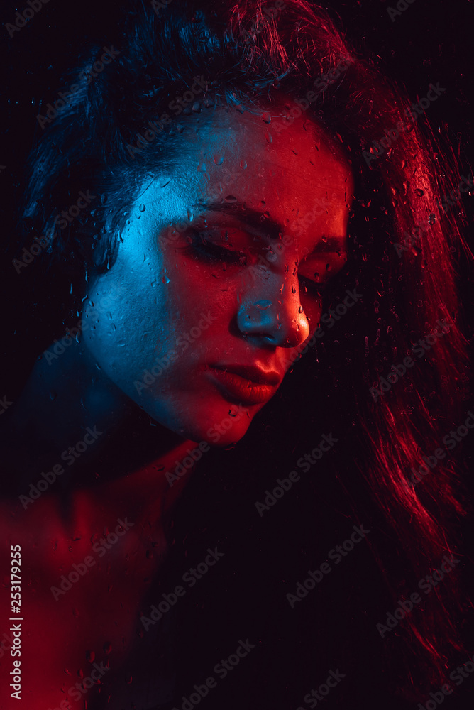 Portrait of sad young girl with red blue lighting behind glass with raindrops