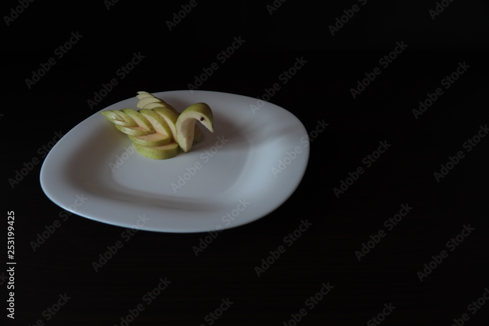 Table setting. The figure of a Swan from a green Apple on a white serving plate, stands on a table with a dark background. Table setting. The figure of a Swan from a green Apple on a white serving plate, stands on a table with a dark background.