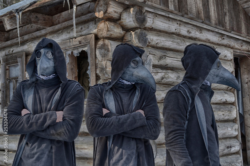 Tree copies (clones) of a man in a mask of a plague doctor and a hood in the forest against the background of an abandoned hut, look in different directions. Something mystical and fabulous. photo