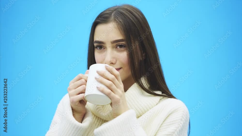 Interestingly, a young woman in a knitted white sweater. Holds a white cup of coffee or tea in his hands. Isolated against the background of a blue wall, studio portrait. Concept of a way of life of Interestingly, a young woman in a knitted white sweater. Holds a white cup of coffee or tea in his hands. Isolated against the background of a blue wall, studio portrait. Concept of a way of life of