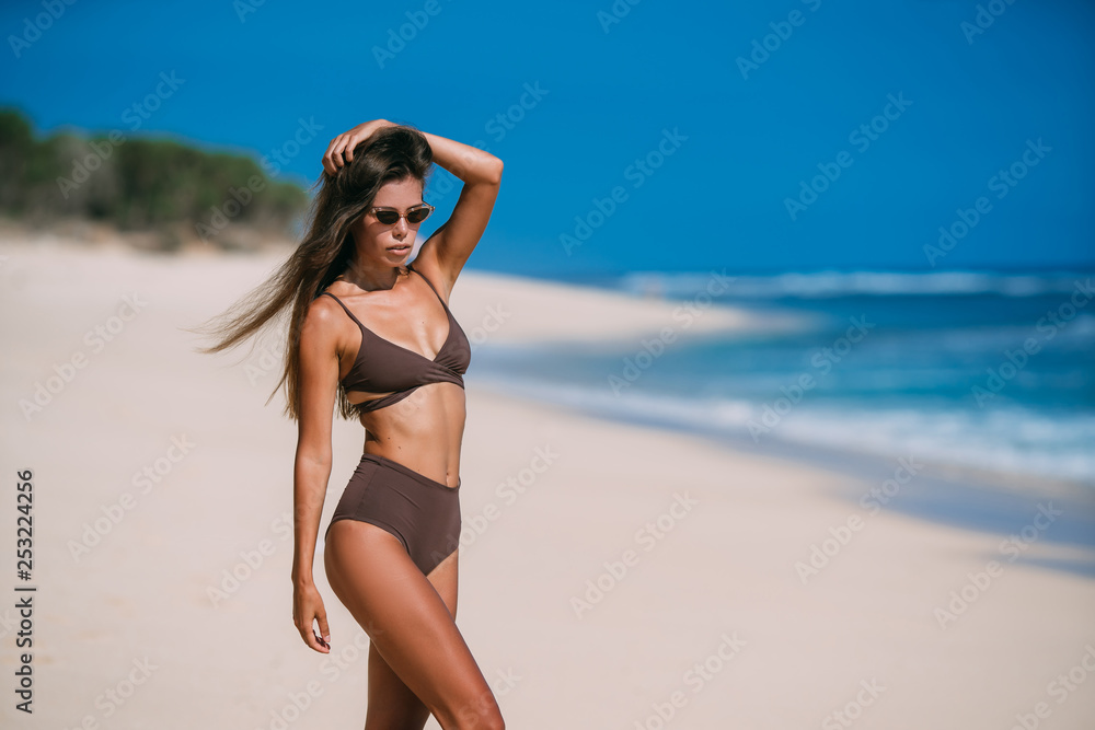 Sexy tanned model with sunglasses in brown swimsuit posing on white sandy beach