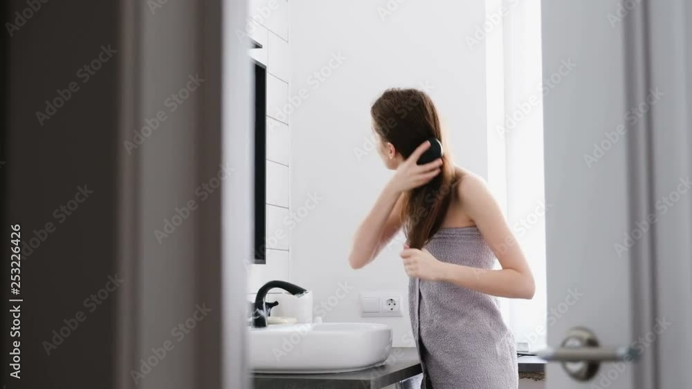 Side view beautiful long-haired young girl combing herself looking in the mirror at her reflection in the bathroom after a shower. Concept of a well-groomed and beautiful body Side view beautiful long-haired young girl combing herself looking in the mirror at her reflection in the bathroom after a shower. Concept of a well-groomed and beautiful body