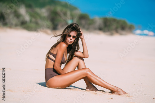 Sexy tanned model with sunglasses in brown swimsuit posing on white sandy beach