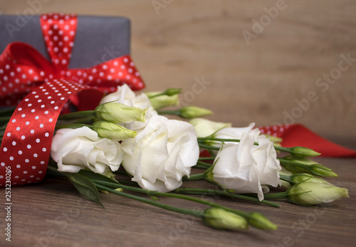 White flowers  eustoma  and a gray gift box with red ribbon in white beans  dots  on a dark wooden background