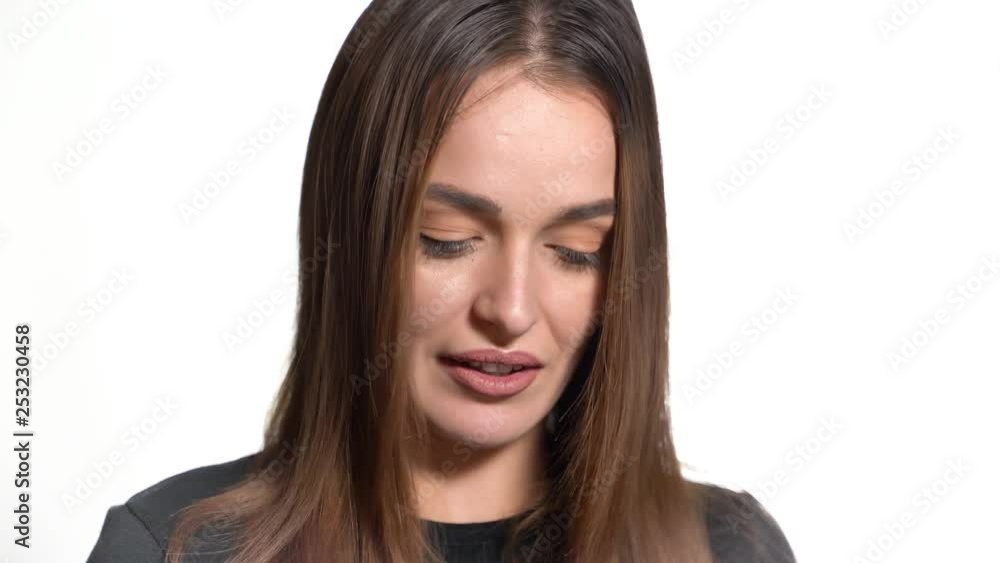 A shy woman with long hair looks down and talks about something on a white background in the studio. Close-up. A shy woman with long hair looks down and talks about something on a white background in the studio. Close-up.