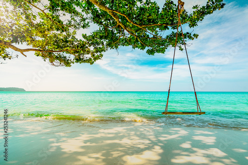 Wooden swings hang from branch of tree at seaside. Emerald green sea water with blue sky and white clouds on summer. Summer vibes. Summer vacation. Empty swings at sand beach with morning sun light . © Artinun