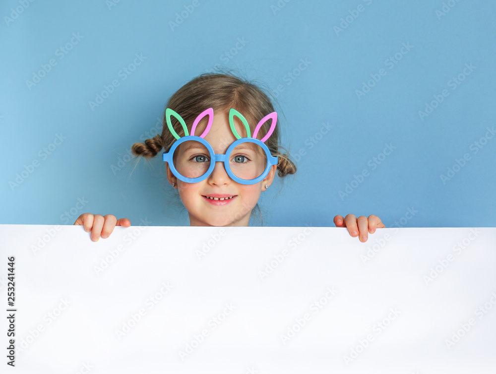 Cute little child wearing bunny ears glasses on Easter day on blue background. Easter girl portrait, funny emotions, surprise. Copyspace for text.
