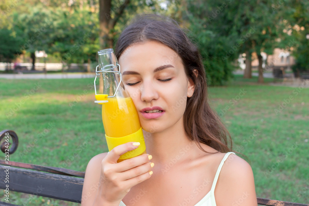 Young woman is tired of hot weather. Girl is holding a cold bottle of juice near forehead Young woman is tired of hot weather. Girl is holding a cold bottle of juice near forehead