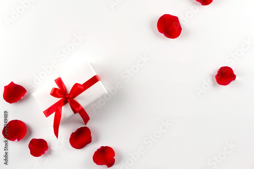 White gift box with red ribbon red velvet rose petals on white background. Flat lay