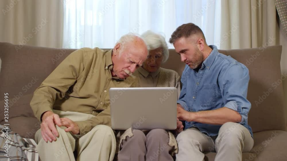 Medium shot of senior couple and their adult son sitting on sofa with laptop and mastering it with the help of their son Medium shot of senior couple and their adult son sitting on sofa with laptop and mastering it with the help of their son