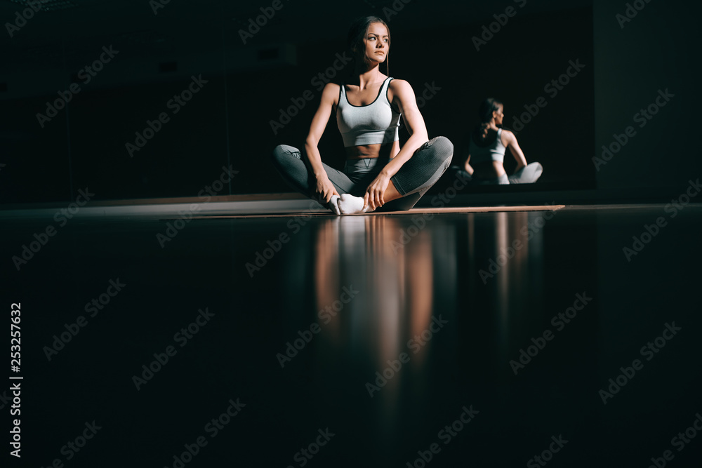 Young Caucasian brunette sitting on the mat in dark and looking away. Fitness studio interior, in background mirror.