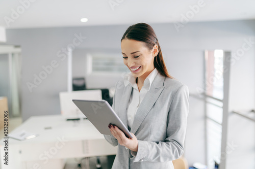 Smiling Caucasian female CEO in formal wear and with long brown hair using tablet while standing at office. Small opportunities are often the beginning of great achievements. © dusanpetkovic1