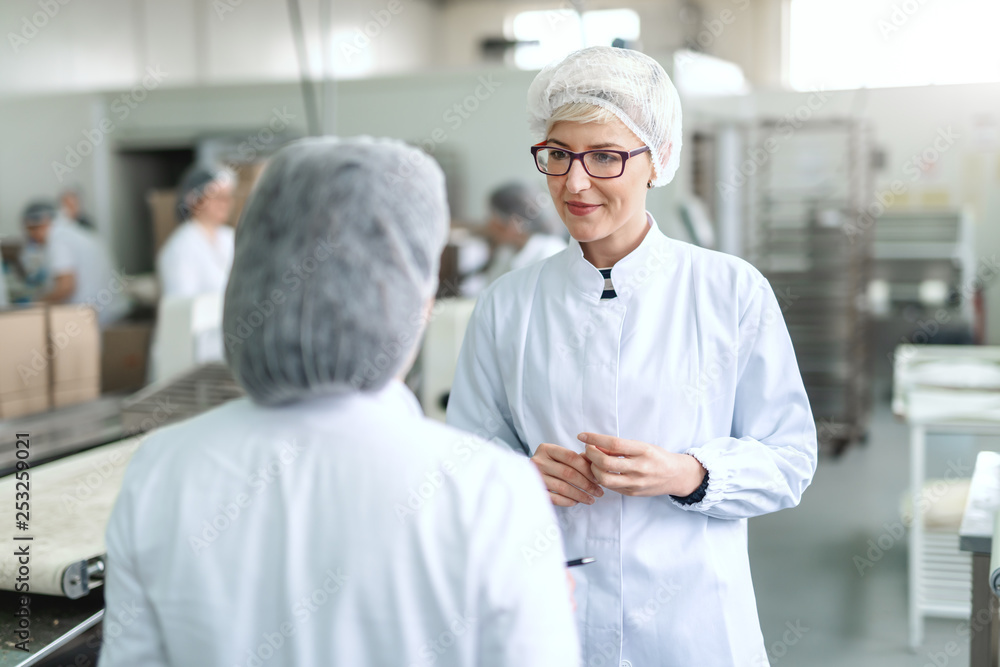 Smiling blonde woman in white sterile uniform and with eyeglasses talking to her boss while standing in food plant.