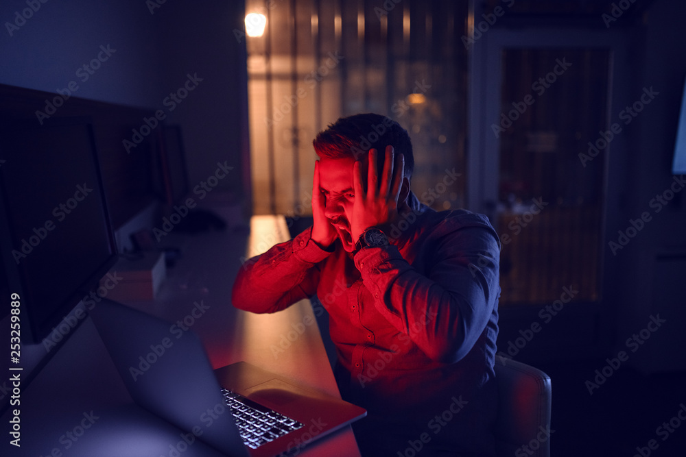 Angry employee with hands on head failing on project. Man looking at laptop and sitting in the office late at night. Angry employee with hands on head failing on project. Man looking at laptop and sitting in the office late at night.
