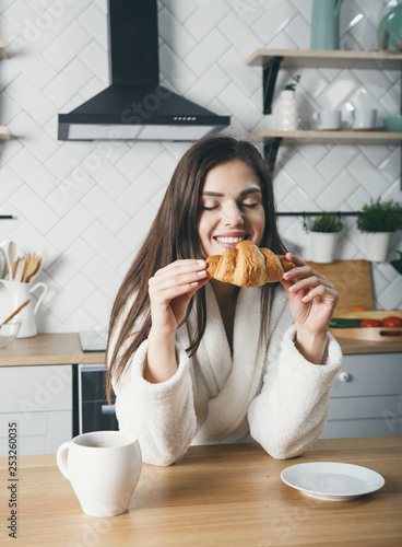 Young brunette girl inhales the aroma of croissant while sitting at the light kitchen