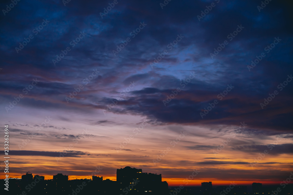 Cityscape with vivid warm dawn. Amazing dramatic blue violet cloudy sky above dark silhouettes of city buildings. Orange sunlight. Atmospheric background of sunrise in overcast weather. Copy space. Cityscape with vivid warm dawn. Amazing dramatic blue violet cloudy sky above dark silhouettes of city buildings. Orange sunlight. Atmospheric background of sunrise in overcast weather. Copy space.