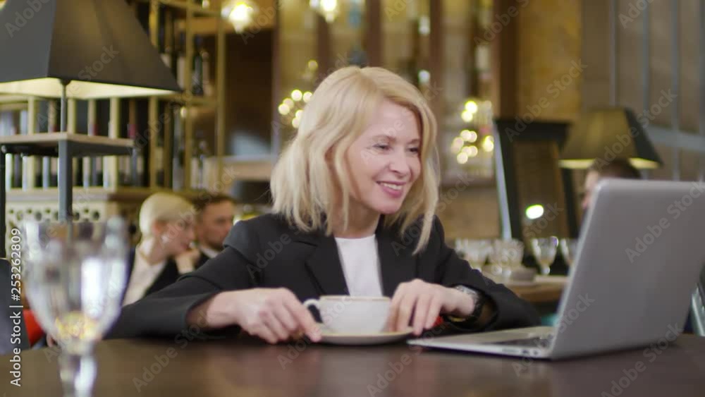 Medium shot of middle-aged Caucasian businesswoman sitting at laptop in café and having online talk Medium shot of middle-aged Caucasian businesswoman sitting at laptop in café and having online talk