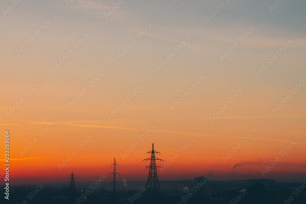 Power lines in city on dawn. Silhouettes of urban buildings among smog on sunrise. Cables of high voltage on warm orange yellow sky. Power industry at sunset. City power supply. Mist urban background. Power lines in city on dawn. Silhouettes of urban buildings among smog on sunrise. Cables of high voltage on warm orange yellow sky. Power industry at sunset. City power supply. Mist urban background.
