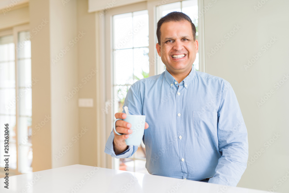 Middle age man drinking coffee in the morning at home with a happy face standing and smiling with a confident smile showing teeth