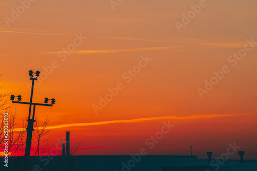 Power lines above roof on dawn. Silhouettes of pile with wires among smog on sunrise. High voltage cables on warm orange yellow sky. Power industry at sunset. City power supply. Mist urban background.