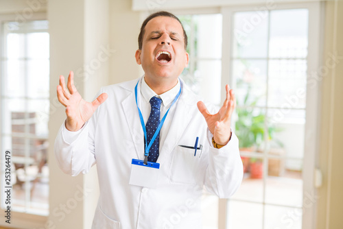 Middle age doctor man wearing medical coat and id card badge over white background celebrating mad and crazy for success with arms raised and closed eyes screaming excited. Winner concept