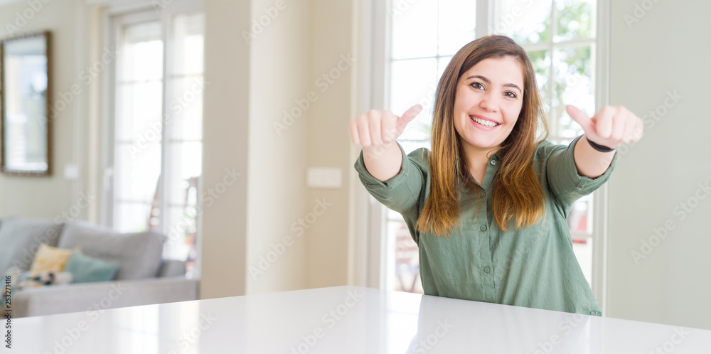 Beautiful young woman at home approving doing positive gesture with hand, thumbs up smiling and happy for success. Looking at the camera, winner gesture. Beautiful young woman at home approving doing positive gesture with hand, thumbs up smiling and happy for success. Looking at the camera, winner gesture.