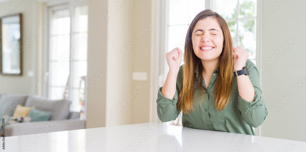 Beautiful young woman at home excited for success with arms raised celebrating victory smiling. Winner concept. Beautiful young woman at home excited for success with arms raised celebrating victory smiling. Winner concept.