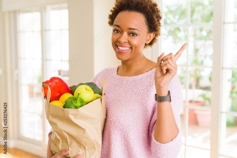 Young african american woman holding paper bag full of fresh groceries very happy pointing with hand and finger to the side Young african american woman holding paper bag full of fresh groceries very happy pointing with hand and finger to the side