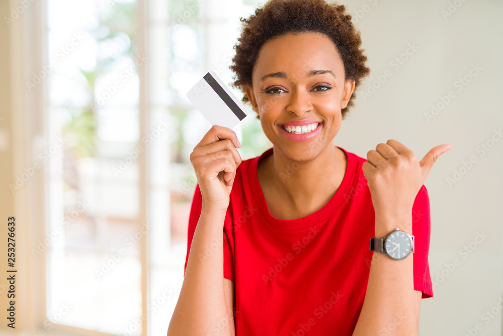 Young african american woman holding credit card pointing and showing with thumb up to the side with happy face smiling Young african american woman holding credit card pointing and showing with thumb up to the side with happy face smiling