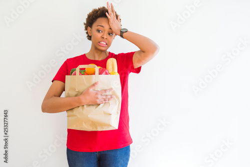 Young african american woman holding paper bag full of fresh groceries stressed with hand on head, shocked with shame and surprise face, angry and frustrated. Fear and upset for mistake.