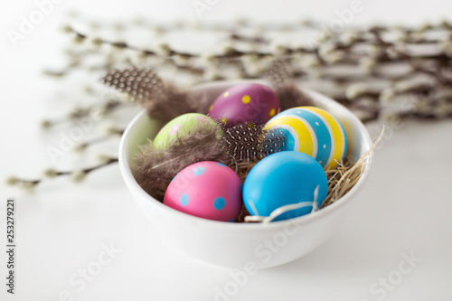 holidays and object concept - colored easter eggs with quail feathers in bowl and pussy willow branches on white background