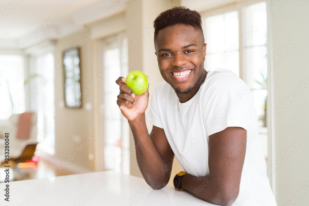 Young african american man eating fresh green apple with a happy face standing and smiling with a confident smile showing teeth Young african american man eating fresh green apple with a happy face standing and smiling with a confident smile showing teeth